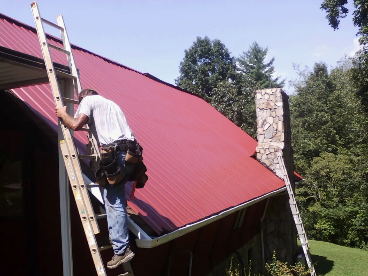 Roofer on a ladder beside a red metal roof and stone chimney — Soffit & Fascia Repair by Roofing Brothers in Pryor